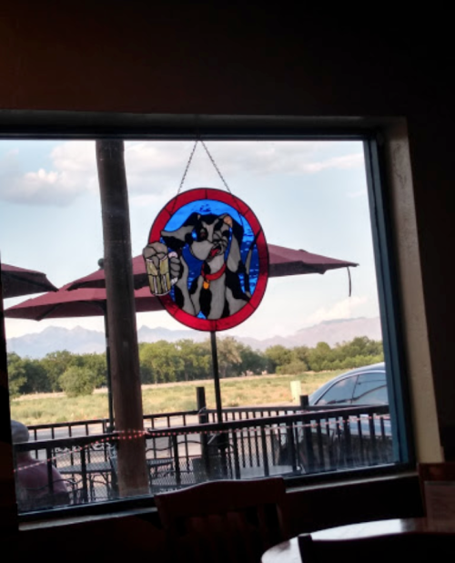 Stained glass window featuring a dog holding a beer, with mountains and umbrellas visible outside.