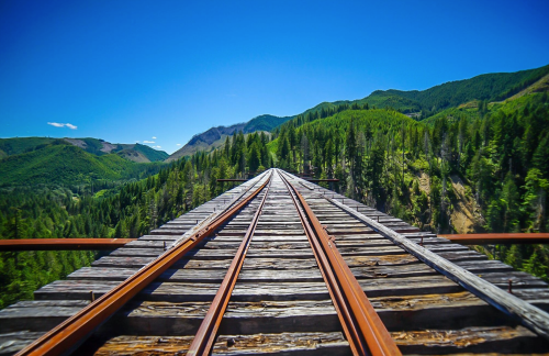 A view of railway tracks stretching into the distance, surrounded by lush green mountains and a clear blue sky.