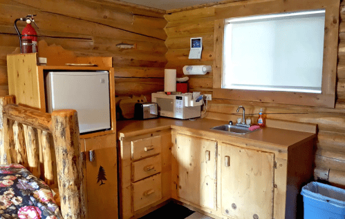 Cozy log cabin kitchen with a small fridge, microwave, sink, and wooden cabinets. Natural wood walls and a floral bedspread.