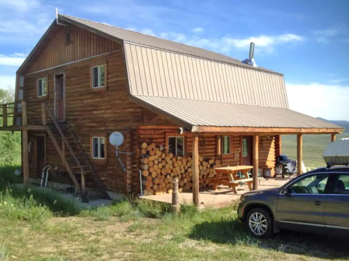 A rustic wooden cabin with a metal roof, stacked firewood, and a car parked nearby, set against a clear blue sky.