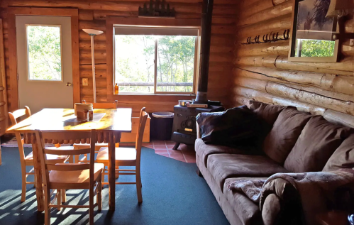 Cozy log cabin interior with a dining table, chairs, a sofa, and a wood stove, surrounded by large windows.