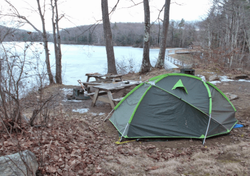 A green tent set up by a lake, surrounded by trees and a picnic area with a table, in early spring.