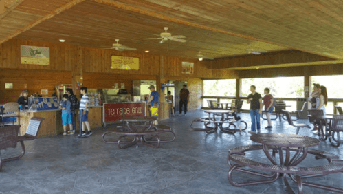 A spacious indoor area with picnic tables, a service counter, and people socializing in a rustic setting.