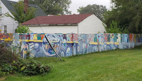 A colorful mural depicting various scenes and patterns on a fence, surrounded by grass and trees.