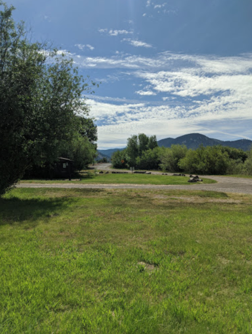 A scenic view of a grassy area with trees, mountains in the background, and a clear blue sky with scattered clouds.