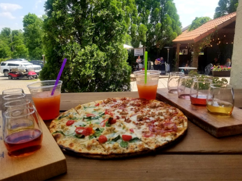 A pizza topped with tomatoes and greens, alongside colorful drinks and tasting flights, set outdoors with greenery.