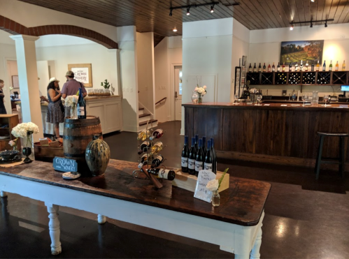 A cozy wine tasting room with a wooden bar, wine bottles, and a rustic table adorned with flowers and a barrel.