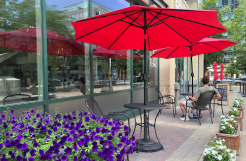 A café patio with red umbrellas, a man sitting at a table, and purple flowers in planters nearby.