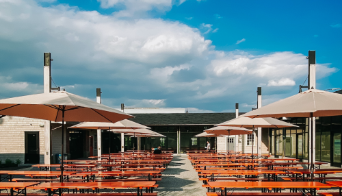 Outdoor dining area with wooden tables, umbrellas, and a blue sky with clouds in the background.