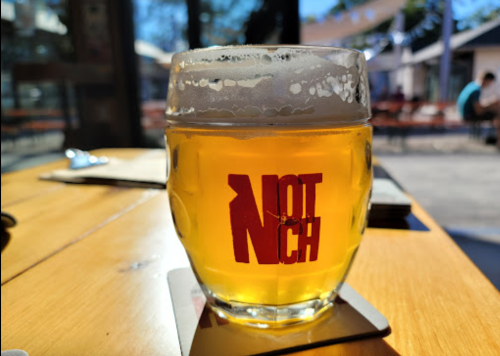 A glass of beer with a frothy top, featuring a red "NOTCH" logo, sits on a wooden table in a sunny outdoor setting.