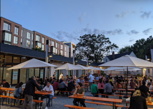 A lively outdoor dining area with people seated at long tables under umbrellas, surrounded by modern buildings at dusk.