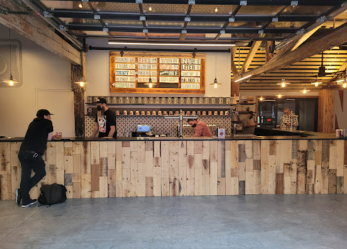 A modern café interior with a wooden counter, two baristas, and a wall of decorative cups and signs.
