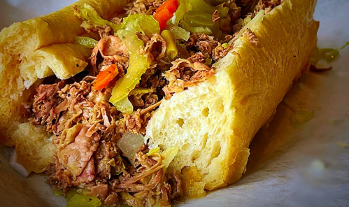 A close-up of a sandwich filled with shredded meat, pickles, and peppers, served in a soft, toasted bun.