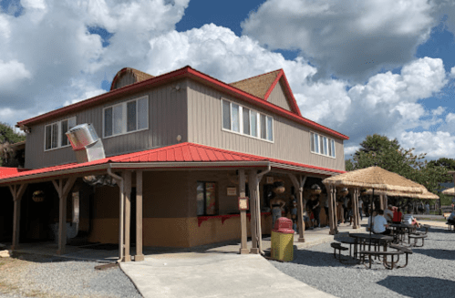 A two-story building with a red roof, surrounded by picnic tables and a thatched-roof structure under a cloudy sky.