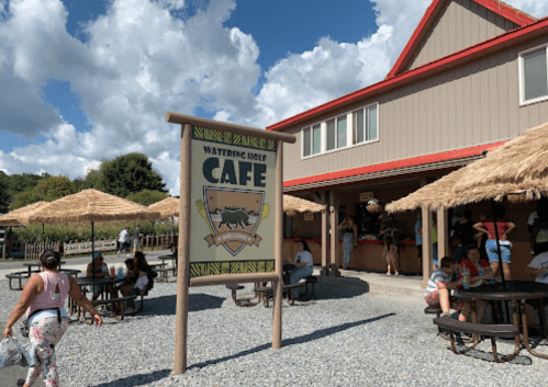 A café sign in front of a building with thatched umbrellas, surrounded by people enjoying the outdoor seating.