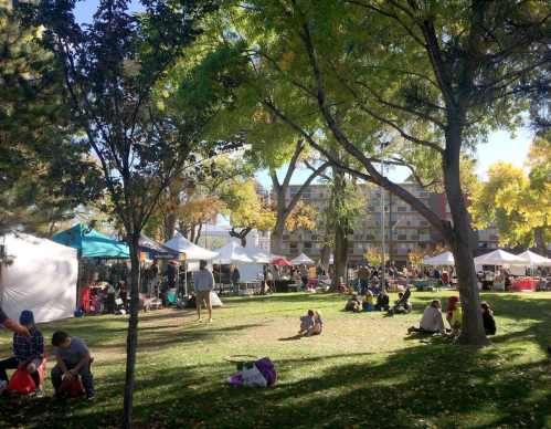 A vibrant outdoor market scene with tents, trees, and people relaxing on the grass under a sunny sky.