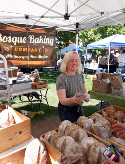 A smiling vendor at a market stands behind a table of freshly baked bread, with a sign for Bosque Baking Company in the background.