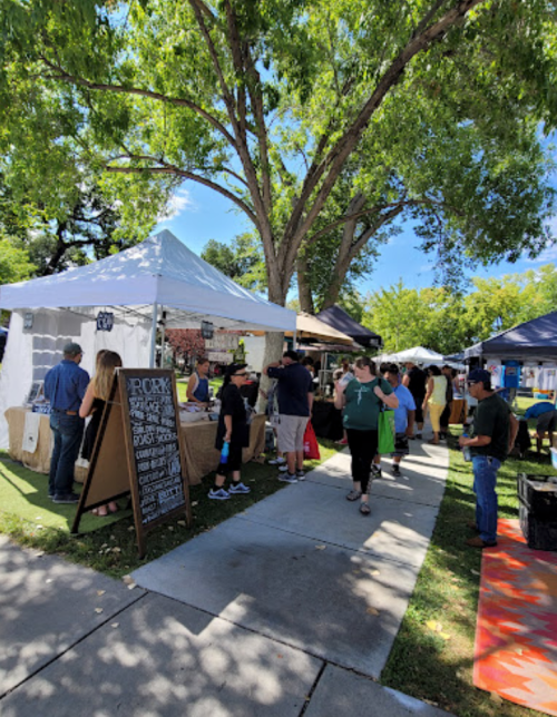 A bustling outdoor market with vendors under tents, people browsing, and trees providing shade on a sunny day.