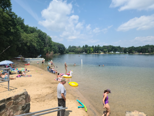 A sunny beach scene with people swimming, lounging, and enjoying the water, surrounded by trees and boats in the background.