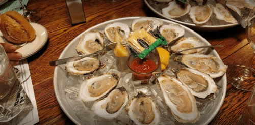 A platter of fresh oysters on ice, garnished with lemon, cocktail sauce, and a small packet of crackers.