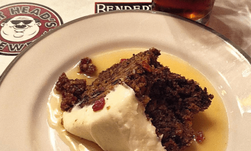 A slice of rich cake topped with ice cream, drizzled with sauce, served on a plate with a drink in the background.