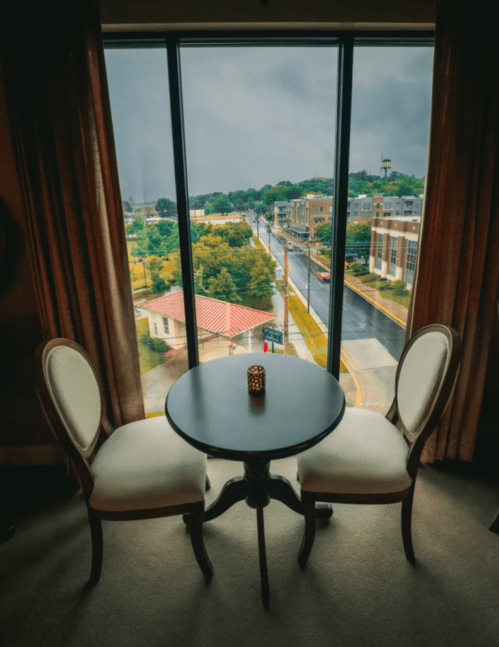 A cozy table with two chairs by a large window overlooking a rainy street and cityscape.