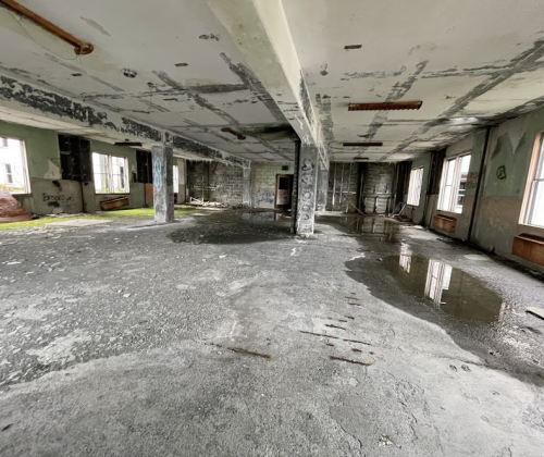 Abandoned interior of a building with cracked walls, puddles on the floor, and large windows letting in natural light.