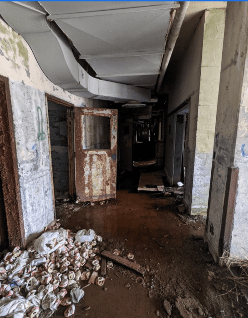 A dimly lit, abandoned hallway with rusted doors and debris, including crumpled cans and scattered trash on the floor.