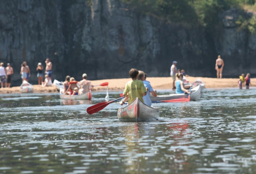 Two people in a canoe paddle on a calm river, with a sandy beach and other people in the background enjoying the sun.