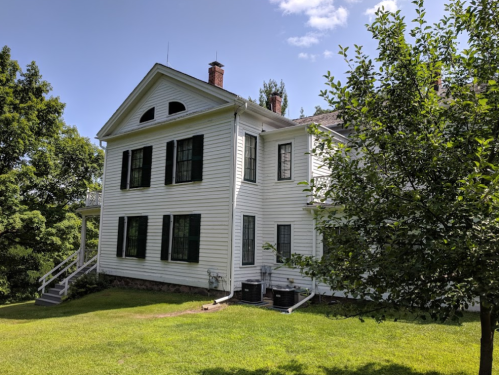 A white two-story house with green shutters, surrounded by trees and a grassy lawn under a blue sky.