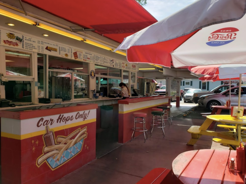 A colorful outdoor diner with red umbrellas, a service counter, and picnic tables in a sunny setting.