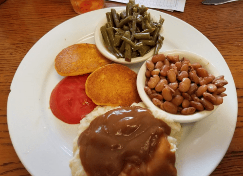 A plate with cornbread, green beans, pinto beans, sliced tomatoes, and a biscuit topped with gravy.