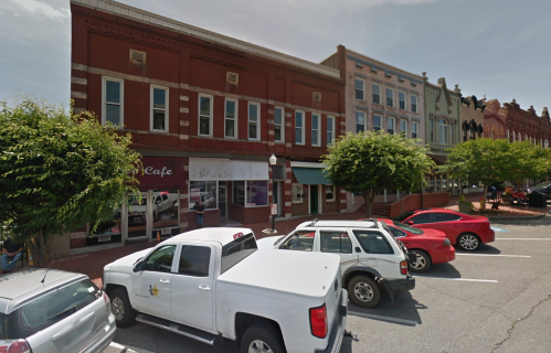 A street view of a historic downtown area with parked cars and trees lining the sidewalk.