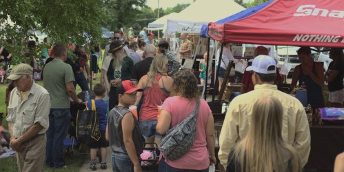 A busy outdoor market scene with people browsing stalls and tents, enjoying a sunny day.