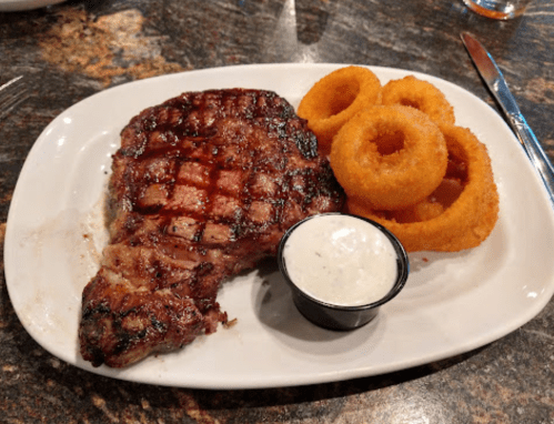 A grilled steak on a white plate, accompanied by onion rings and a small cup of dipping sauce.