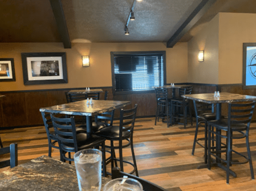 Interior of a restaurant with empty tables, black chairs, and wooden flooring, featuring dim lighting and framed photos.