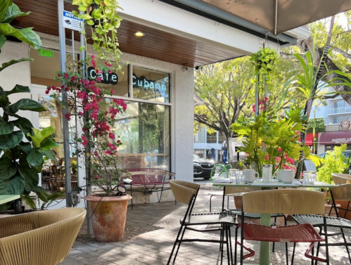 Outdoor café scene with colorful plants, tables, and chairs under a shaded patio. Bright and inviting atmosphere.