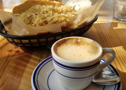 A small cup of coffee on a plate next to a basket of golden, shredded food on a wooden table.