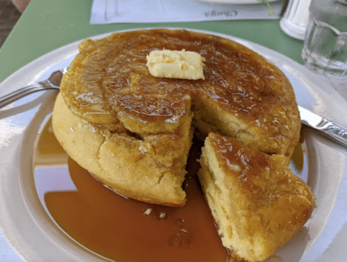 A thick, golden pancake topped with a pat of butter and drizzled with syrup, with a slice cut out on a white plate.