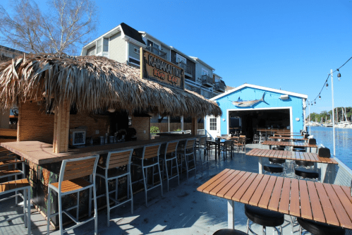 A waterfront bar with a thatched roof, wooden tables, and a blue building in the background, under a clear blue sky.