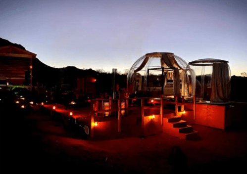 A cozy outdoor setup featuring transparent domes, illuminated pathways, and a scenic mountain backdrop at dusk.