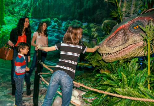 A family interacts with a dinosaur exhibit in a lush, green environment, with one child reaching out to touch the dinosaur.