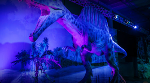 A large, colorful dinosaur sculpture in a dimly lit exhibit, surrounded by palm trees and vibrant lighting.
