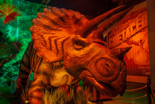 A large, colorful dinosaur model resembling a Triceratops in a themed exhibit with vibrant lighting and foliage.