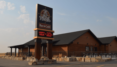 Sign for 3 North Bar & Grill with a stone base, featuring a happy hour promotion, set against a rustic building.