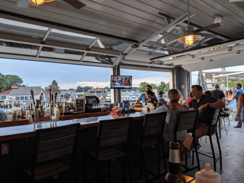 A bar with patrons enjoying drinks, large windows showing a waterfront view, and a TV displaying a sports game.