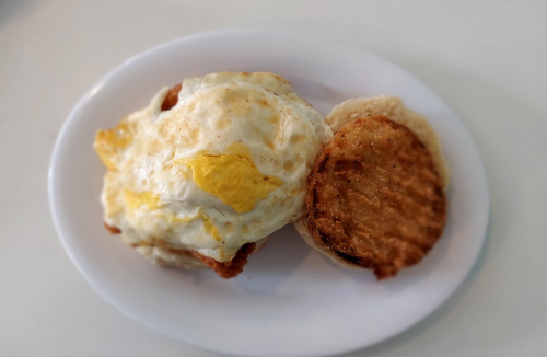 A breakfast sandwich with a fried egg and a golden-brown sausage patty on a toasted biscuit, served on a white plate.