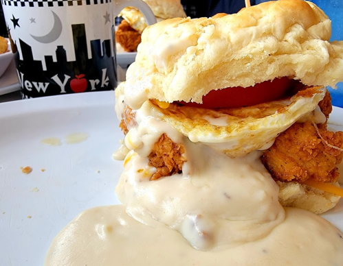 A hearty sandwich with fried chicken, a fried egg, tomato, and creamy sauce, served on a biscuit with a coffee mug in the background.
