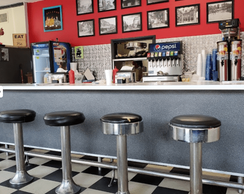 A retro diner counter with black stools, soda dispensers, and vintage photos on the red wall.