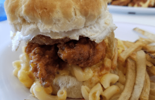 A biscuit sandwich with fried chicken, macaroni and cheese, and a fried egg, served with French fries on the side.